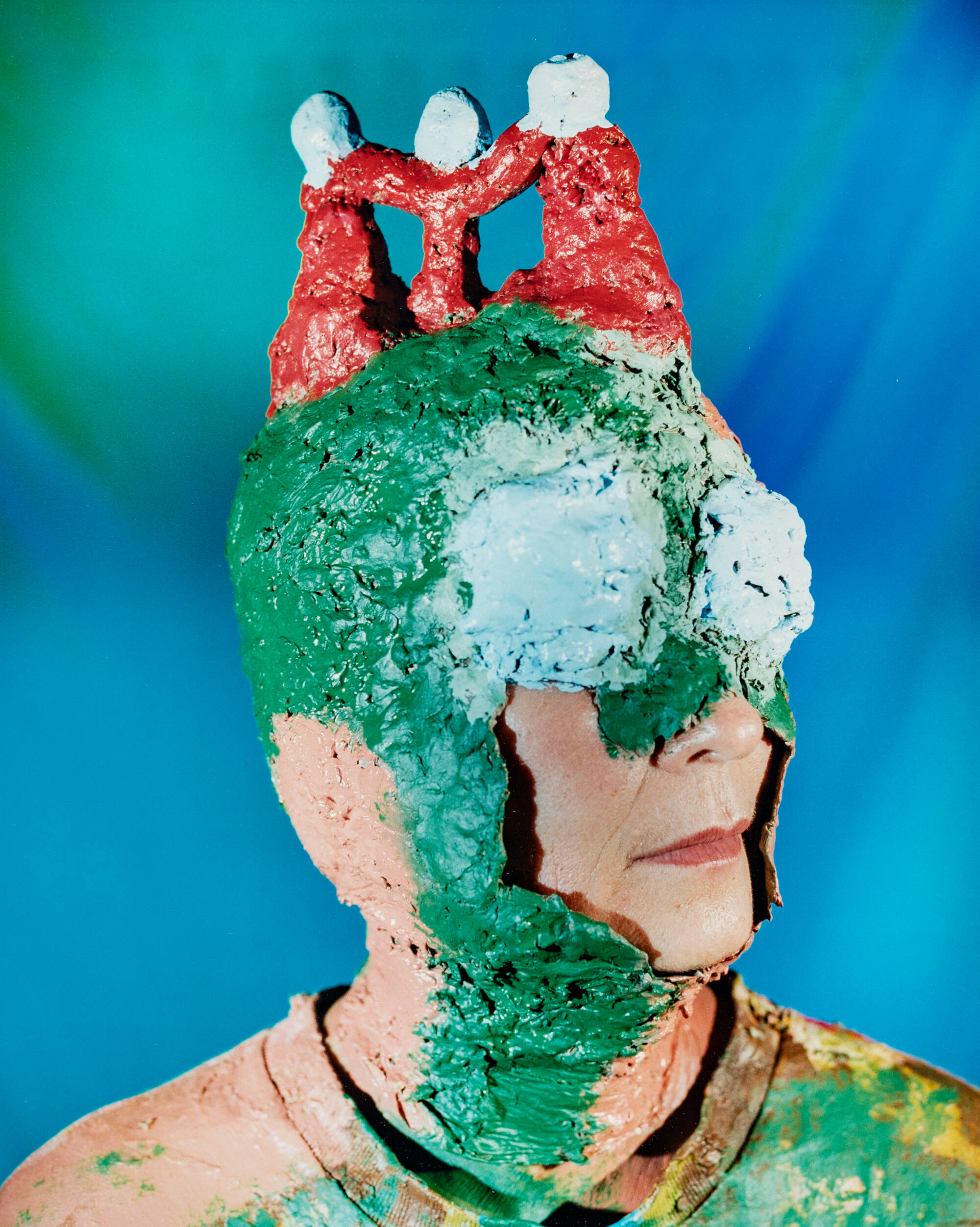 Colorful photograph of a person wearing a mask that covers everything but their nose and mouth. Mask is bright green and red, background is bright blue.